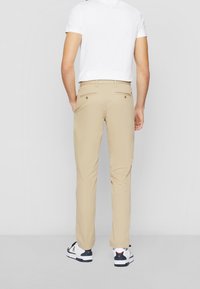 Pantalons beiges, à jambe droite, en tissu léger avec deux poches arrière comportant des boutons dorés. Porté avec une chemise blanche à manches courtes et des baskets.