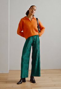 Suéter de punto naranja con escote en V y textura de canalé, combinado con pantalones de pierna ancha verdes con pliegues y botas negras hasta el tobillo.