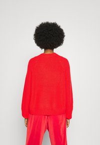 Rode gebreide trui met een ronde hals, geribde manchetten en een losse pasvorm. De textuur is chunky en de achterkant is eenvoudig zonder versieringen.