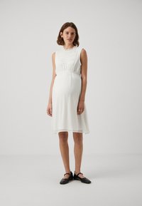 Robe blanche sans manches avec un motif texturé et un col en volants, froncée sous la poitrine, tombant juste au-dessus des genoux. Chaussures plates noires.
