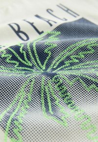 Gros plan sur un tissu imprimé de palmiers à pois bleus et orné de détails de feuilles de palmier vertes brodées. Le mot "BEACH" est partiellement visible.