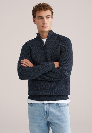 Suéter texturizado azul marino con cuello de media cremallera, tejido de punto y puños acanalados. Lleva puesto sobre una camisa blanca, combinado con jeans de color azul claro.