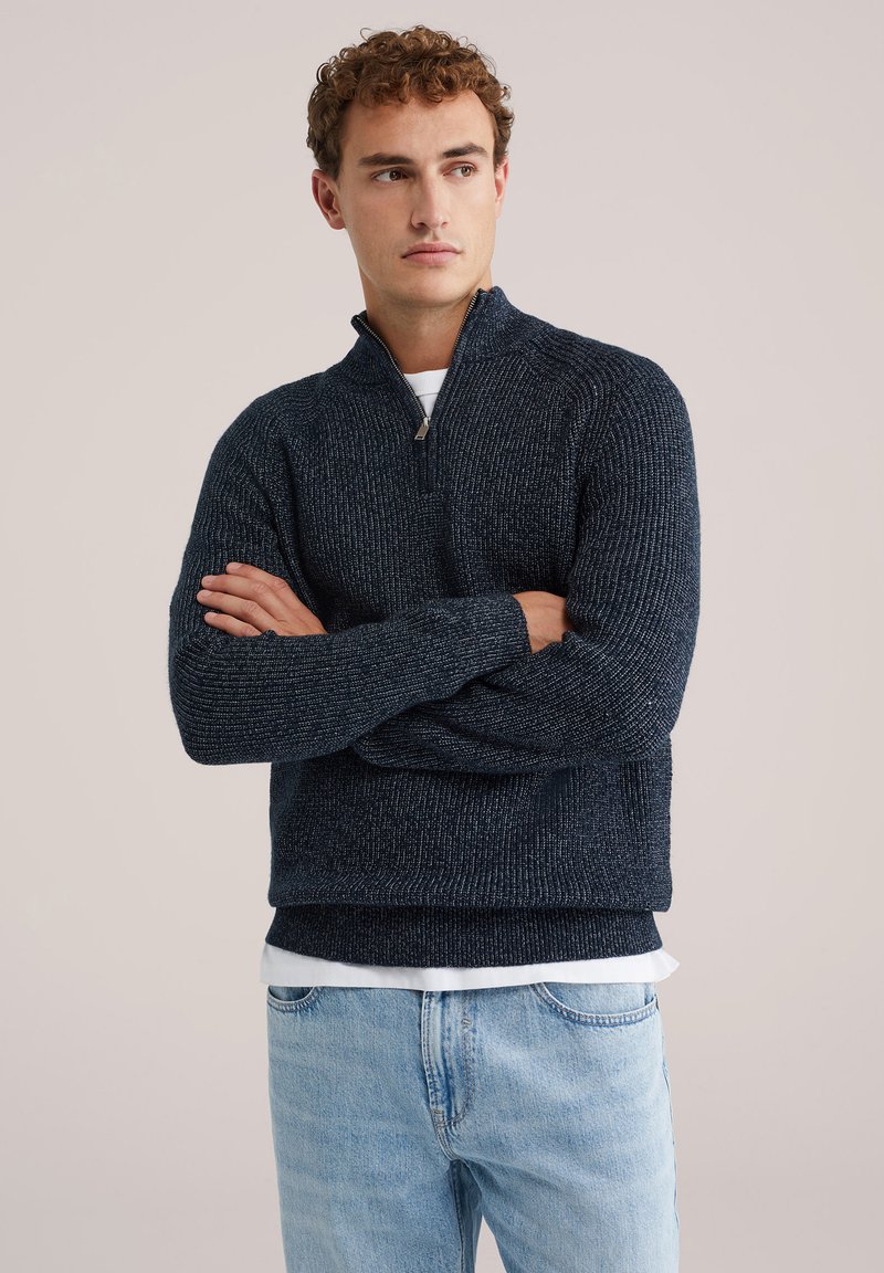 Texturēts tumši zils puloveris ar ceturtdaļas zibeni, adīta auduma, un ribotām piedurknēm. Uzvilkts virs baltas krekla, saskaņots ar gaiši ziliem džinsiem.