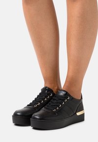 Black platform sneakers made of textured material, featuring gold eyelets, black laces, and a black sole with a gold accent.