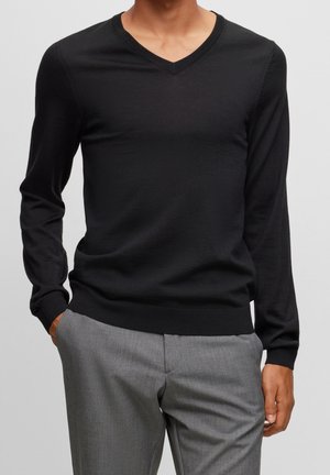 Man draagt een nauwsluitende zwarte trui met V-hals en lange mouwen en grijze broek, handen in de zakken, tegen een lichtgrijze achtergrond.