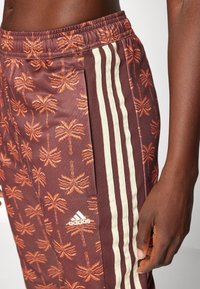 Maroon joggerbyxor med palmträdsmönster, vit adidas-logotyp och tre vita sidostråk. Tyget har en slät textur.