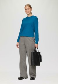 Turquoise lange mouwen trui gecombineerd met grijze wijde broek; vasthoudend een gestructureerde zwarte schoudertas met een klein ritsvak. Zwarte schoenen.