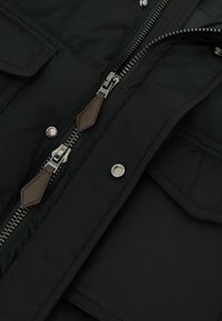 Close-up van een zwart jack met dubbele metalen ritsen met bruine leren trekker, drukknopen en klepzakken op een stoffen textuur.