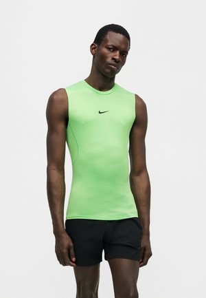 Atletyczny mężczyzna w obcisłej, bezrękawnikowej neonowozielonej koszulce Nike i czarnych spodenkach, stojący na jednolitym jasnym tle.