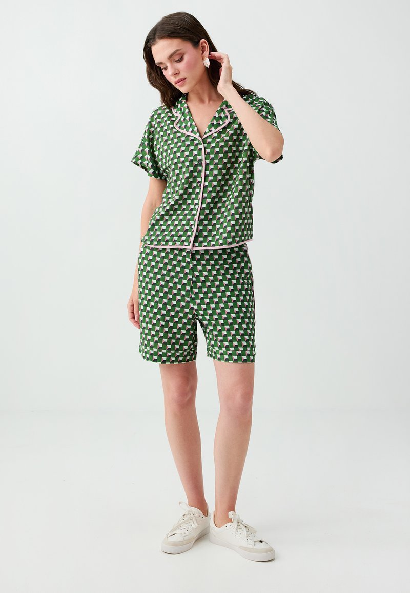 Femme portant une chemise boutonnée à manches courtes à motif vert et un short assorti, avec des baskets blanches, debout dans un espace clair et neutre.