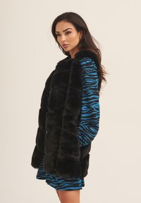 Black faux fur vest with a plush texture, featuring a high collar and structured shape. Paired with a blue and black striped dress.