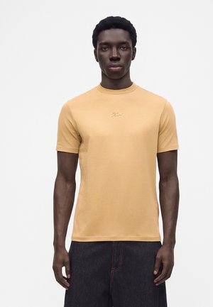 Junger Mann trägt beige kurzärmeliges T-Shirt und dunkle Hose, steht frontal vor weißem Hintergrund.