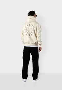 Cream hoodie with a spiral graphic in pink and black text on the back, paired with black pants and white sneakers. Smooth texture.