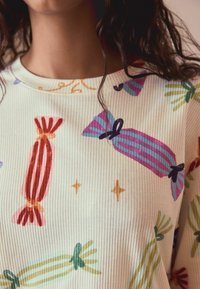 Hvid ribbet sweater med et gentagende mønster af farverige slik og stjerner, med accenter i pink, grøn, lilla og orange.