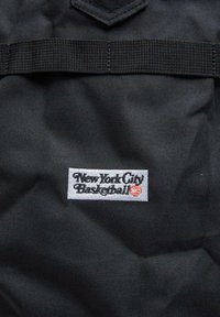Black fabric backpack with a prominent white rectangular label reading "New York City Basketball," featuring black and orange text details.