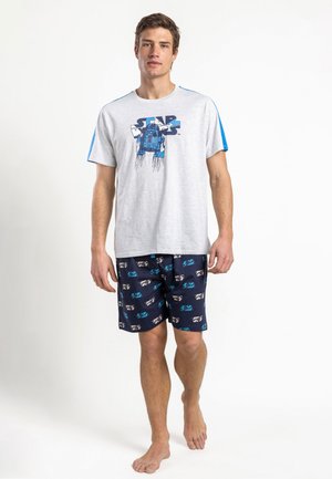 Hombre con camiseta de Star Wars de color gris claro con rayas azules en las mangas y pantalones cortos con motivo de Star Wars en azul oscuro, de pie descalzo.