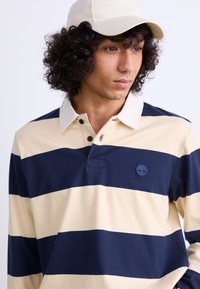 Polo majica s dolgimi rokavi v kremnih in mornarsko modrih črtah, z gumbi na zankah in logotipom na prsih. Kapa v kremni barvi.