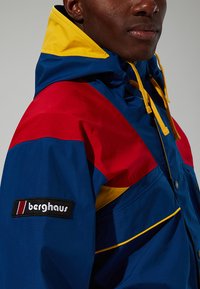 Bunter Windbreaker mit blauen, roten und gelben Paneelen. Verfügt über eine Vordertasche und ein schwarzes "Berghaus"-Logo am Ärmel.