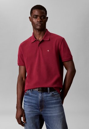 Mann trägt ein rotes kurzärmeliges Polohemd mit kleinem weißem Logo und blauen Jeans vor einem einfarbigen grauen Hintergrund.