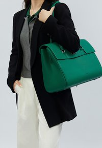 Grøn tekstureret håndtaske med en struktureret form, sølv hardware og en topklap, båret af en person i en sort jakke og grå skjorte.