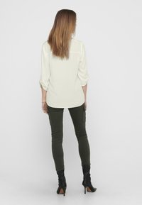 Camisa blanca de manga larga con puños remangados, combinada con pantalones cargo ajustados verde oliva y botas negras de tacón alto. Textura suave, ajuste casual.