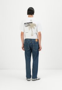 Bărbat îmbrăcat cu un tricou alb cu grafică, având un palmier și textul "Riverside County," blugi albaștri și adidași albi, stând cu fața spre un perete alb.