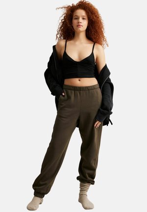 Ung kvinde med krøllet rødt hår iført en sort crop top, løse mørke olivengrønne joggingbukser, grå sokker og en sort jakke hængende over skuldrene.