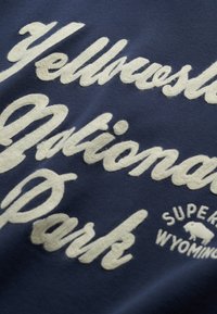 Tela de color azul marino con bordado texturizado en blanco que dice "Parque Nacional Yellowstone" y "Super Wyoming" junto a un gráfico de un bisonte.