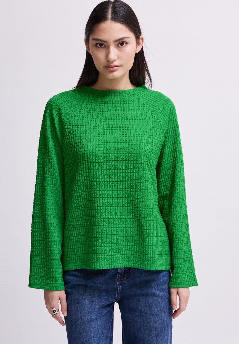 Maglia verde texturizzata a maniche lunghe con forma quadrata e maniche raglan, abbinata a jeans in denim blu. Il tessuto presenta un motivo a griglia.