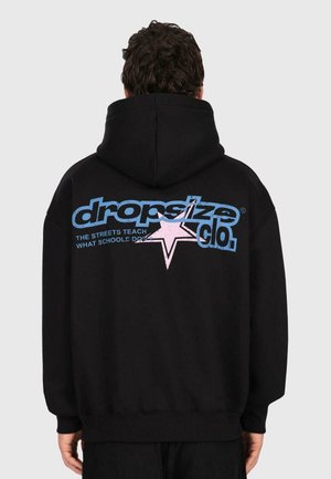 Un homme portant un sweat à capuche noir avec un grand texte "dropsize clo." en bleu et rose et un graphique d'étoile dans le dos, se tenant devant un fond uni.