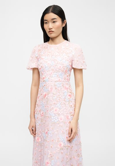Νεαρή γυναίκα με μακριά σκούρα μαλλιά, που φοράει ένα ανοιχτό ροζ φόρεμα με κοντά μανίκια και παστέλ κεντήματα με λουλούδια.