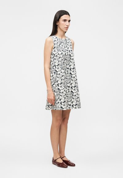 Γυναίκα που φοράει αμάνικο ασπρόμαυρο φόρεμα με λουλούδια και καφέ παπούτσια Mary Jane, στέκεται μπροστά σε απλό ανοιχτό γκρι φόντο.