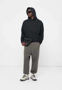 Schwarzer Kapuzenpullover mit Kängurutasche, kombiniert mit lockeren olivgrünen Hosen. Schwarze Sonnenbrille und graue strukturierten Sneakers.