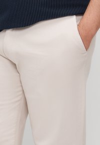 Beige cotton trousers with side pockets, smooth texture, tailored fit, and minimal design. Visible navy ribbed garment above.