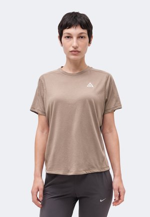 Kobieta z krótkimi ciemnymi włosami nosi beżowy t-shirt z logo ACG i czarne legginsy Nike, stoi na jednolitym jasnym tle.