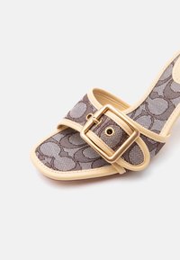 Πέδιλο με ανοιχτή μύτη, μπεζ περιθώριο, καφέ υφασμάτινο λουρί με σχέδιο και μεγάλη τετράγωνη χρυσή αγκράφα σε ανοιχτό φόντο.