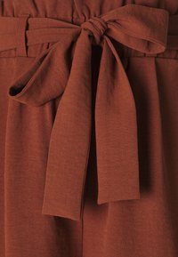 Ceinture en tissu de couleur rouille nouée en un nœud papillon, attachée à un pantalon. Le matériau semble doux avec une finition légèrement texturée.
