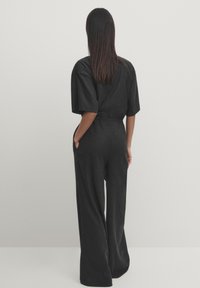 Black wide-leg jumpsuit with short sleeves, featuring a cinched waist and side pockets. Soft fabric with a subtle texture.