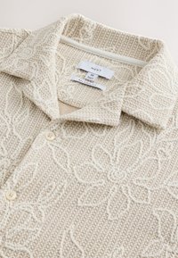 Chemise en maille texturée beige et crème avec motifs floraux, boutonnée devant, col ouvert, étiquette de la marque "Next" taille moyenne, coupe décontractée.