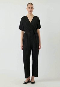 Macacão preto com pregas, design envolvente, mangas curtas e cintura ajustável, apresentando pernas retas e uma textura suave.