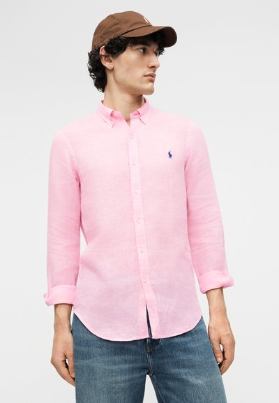 Vīrietis, tērpts gaiši rozā pogātu kreklu ar uzruļētām piedurknēm, zilos džinsos un brūnā cepurē, raugās sāniski uz vienkārša fona.