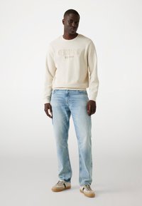 Sudadera crema con el logo en relieve "GUESS EST.1981", combinada con unos jeans de mezclilla azul claro y zapatillas beige con acentos verdes.