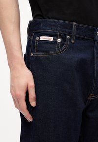 Jeans de denim azul oscuro con costuras amarillas, que cuentan con una etiqueta de Calvin Klein en el bolsillo, diseño ajustado y detalles de metal.