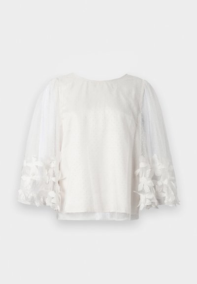 Blusa blanca transparente con lunares, mangas largas abullonadas y puños bordados con flores, expuesta sobre fondo blanco liso.