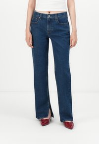Jean en denim bleu foncé avec une coupe droite, dotée de poches avant, d'une fermeture à bouton et de détails de couture subtils ; accompagnée de chaussures rouges.