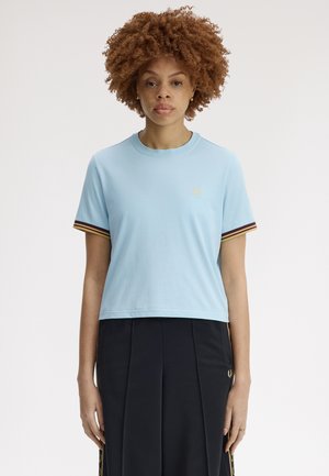 Kvinde med krøllet hår iført en lyseblå kortærmet skjorte og sort nederdel, som står foran en ensfarvet hvid baggrund.