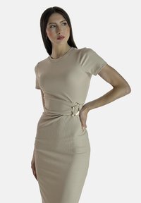 Een vrouw met lang donker haar poseert zelfverzekerd in een aansluitende beige jurk met een gouden riemdetail, wat haar stijlvolle look benadrukt.
