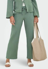 Grønt teksturert to-delt antrekk med justerbar midje, vide bukser og jakke med knyting foran, kombinert med en beige totebag og sandaler.