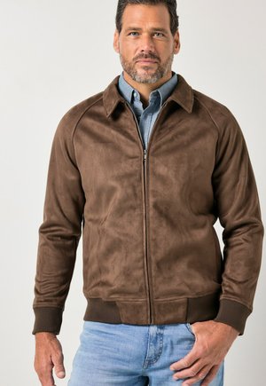 Mand iført brun ruskind zip-up jakke over blå skjorte med knapper og lyseblå jeans, stående foran en neutral baggrund.