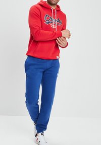 Czerwona bluza z kapturem z logo z przodu, białe sznurki do ściągania i niebieskie spodnie dresowe. Posiada ściągacze przy mankietach i w pasie, a także białe sneakersy.